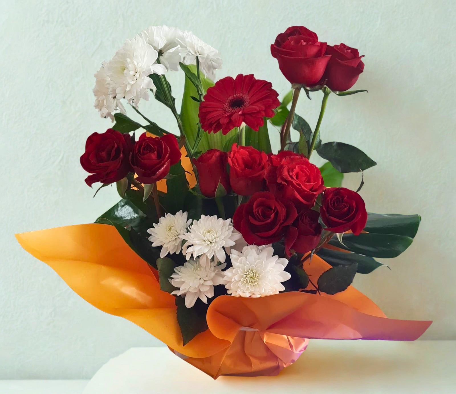Elegant bouquet featuring red and white flowers with lush green foliage, beautifully wrapped in vibrant orange paper, perfect for gifting on special occasions.